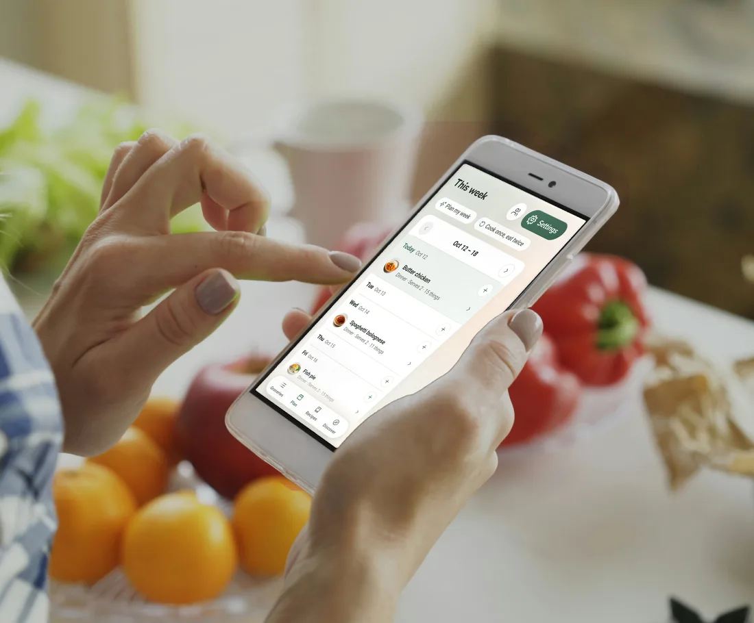 Hands holding a phone at a kitchen table set with oranges, apples and red peppers, the phone showing this week in Fridg: butter chicken on Monday, spaghetti bolognese on Wednesday and fish pie on Friday